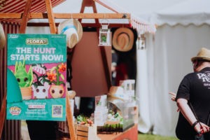 Funko Flora Pops Bunte Werbetafel für das Funko "Flora"-Event am Elbenwald Festival, die auf die bevorstehende Veröffentlichung am 24. August hinweist. Im Hintergrund sind verschiedene dekorative Pflanzen und Merchandise-Artikel zu sehen, während ein Besucher interessiert schaut.