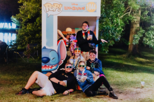 Sechs fröhliche Festivalbesucher posieren vor einem Disney Lilo & Stitch Fotopunkt beim Elbenwald Festival. Sie tragen bunte Sonnenbrillen und Blumenketten, während sie Getränke halten und eine entspannte Atmosphäre genießen.