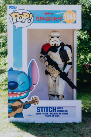 Ein Stormtrooper in einer Vitrine, umgeben von einem bunten Hintergrund mit Stitch aus Disney's "Lilo & Stitch". Die Figur hält ein Gewehr und steht in einer kreativen Pose, die die Verbindung von Popkultur und Fandom auf dem Elbenwald Festival zeigt.