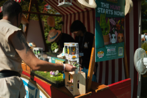 Ein Besucher am Stand beim Elbenwald Festival, der eine Figur aus einer Sammlung auswählt. Im Hintergrund sind weitere Figuren und ein Werbeplakat für eine bevorstehende Produktveröffentlichung zu sehen. Bunte Festivalatmosphäre und kreative Stände prägen das Bild.