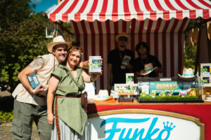 Funko Flora Pops Zwei Festivalbesucher posieren vor einem bunten Funko-Stand auf dem Elbenwald Festival. Sie halten eine Funko-Pop-Figur in der Hand, während im Hintergrund weitere Figuren und Merchandise-Artikel ausgestellt sind. Strahlende Gesichter und festliche Atmosphäre.