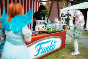 Funko Flora Pops Besucher des Elbenwald Festivals am Funko-Stand, der verschiedene Sammlerstücke und Merchandise anbietet. Im Vordergrund sind zwei verkleidete Personen zu sehen, eine mit orangefarbenen Haaren und die andere mit langen, pinken Haaren.