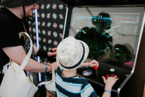Gamesroom by Nerdistan Vater und Sohn spielen an einem Retro-Arcade-Automaten während des Elbenwald Festivals, umgeben von bunten Grafiken. Die Szene zeigt die Freude an Videospielen und die familiäre Bindung in einer nerdigen Festivalatmosphäre.