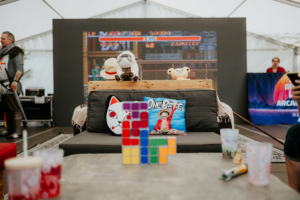 Gamesroom by Nerdistan Bunte Kissen und Spielzeugfiguren auf einer Couch im Elbenwald Festival-Zelt, im Hintergrund ein Retro-Arcade-Spiel auf einem Bildschirm. Auf dem Tisch stehen Getränke und ein Rubik's Cube, der die nerdige Atmosphäre des Festivals unterstreicht.