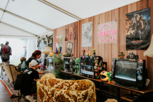 Gamesroom by Nerdistan Gemütlicher Gaming-Bereich auf dem Elbenwald Festival, mit Retro-Fernsehern, bunten Figuren und einem einladenden Interieur. Besucher spielen an Konsolen und genießen die nerdige Atmosphäre in einem Zelt voller kultureller Anspielungen.