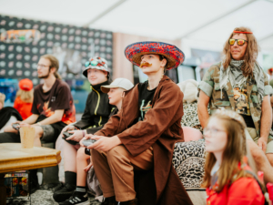 Gamesroom by Nerdistan Eine Gruppe von Festivalbesuchern in kreativen Kostümen sitzt entspannt auf einem Sofa und spielt Videospiele. Die Szene zeigt die lebendige Atmosphäre des Elbenwald Festivals, wo Musik und Nerdkultur verschmelzen.