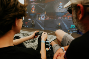 Gamesroom by Nerdistan Zwei Personen spielen gemeinsam ein Videospiel auf einem großen Bildschirm beim Elbenwald Festival. Der Fokus liegt auf dem Spieler mit einem Controller in der Hand, während spannende Spielszenen im Hintergrund zu sehen sind.