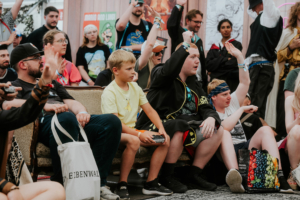 Gamesroom by Nerdistan Teilnehmer des Elbenwald Festivals sitzen auf einer Couch und feuern begeistert eine Veranstaltung an. Ein Junge mit einem Gamecontroller in der Hand sitzt zwischen mehreren Fans, die in Kostümen gekleidet sind und ihre Hände heben. Die Atmosphäre ist lebhaft und enthusiastisch.