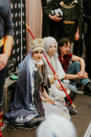 Zwei Cosplayerinnen in aufwendigen Kostümen, eine mit einer königlichen Kopfbedeckung und einem Umhang, die andere in einem weißen Gewand, sitzen auf dem Boden während des Elbenwald Festivals. Im Hintergrund sind weitere Festivalbesucher und Dekorationen zu sehen.