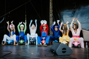 Mario Kart Turnier-Finale Eine Gruppe von Cosplayern in bunten Kostümen, die Charaktere aus einem Videospiel darstellen, sitzt auf kleinen Fahrzeugen auf der Bühne des Elbenwald Festivals. Die Teilnehmer heben die Hände und zeigen ihre Begeisterung für das Event.