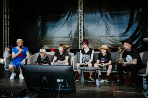 Mario Kart Turnier-Finale Eine Gruppe von fünf Personen, darunter Kinder und Erwachsene, sitzt auf Sofas und spielt Videospiele auf einer Bühne beim Elbenwald Festival. Ein Moderator mit Mikrofon spricht, während die Spieler konzentriert auf den Bildschirm schauen.
