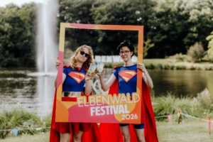 Zwei Teilnehmerinnen in Superheldenkostümen halten ein Fotorahmen-Schild des Elbenwald Festivals 2026. Im Hintergrund ist ein Teich mit einem Springbrunnen zu sehen, umgeben von grüner Natur und bunten Dekorationen.
