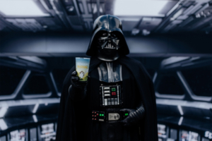 Darth Vader hält ein Getränk mit dem Logo des Elbenwald Festivals in einem futuristischen Setting. Die Atmosphäre verbindet Musik und Nerdkultur, während der ikonische Charakter Fans aus der Popkultur anzieht. Ideal für Festivalbesucher und Cosplay-Enthusiasten.
