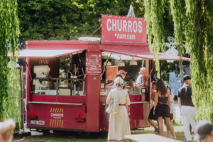Churros-Stand auf dem Elbenwald Festival, umgeben von Besuchern in bunten Kostümen. Der Foodtruck bietet frisch zubereitete Churros und Snacks an, während die Festivalatmosphäre lebendig und einladend ist.
