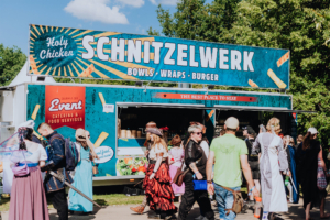Buntes Treiben am Essensstand "Schnitzelwerk" beim Elbenwald Festival, wo Besucher in kreativen Kostümen eine Auswahl an Bowls, Wraps und Burgern genießen. Der Stand bietet eine einladende Atmosphäre für Festivalteilnehmer und Food-Liebhaber.