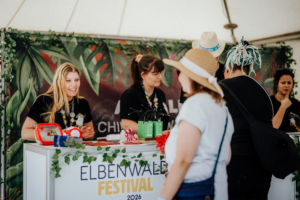 Mitarbeiterinnen am Stand des Elbenwald Festivals 2026, umgeben von Pflanzen-Dekoration. Besucherinnen interagieren mit dem Team, während Merchandise und Informationen angeboten werden. Eine lebendige Atmosphäre, die das nerdige Festivalerlebnis widerspiegelt.
