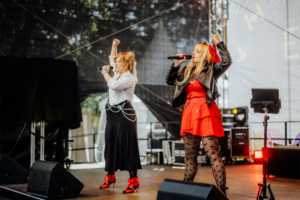 Harmoonics Zwei Sängerinnen performen auf der Bühne des Elbenwald Festivals, gekleidet in auffälligen Outfits. Die Atmosphäre ist lebhaft, während das Publikum die energiegeladene Darbietung genießt. Im Hintergrund sind technische Geräte und Bühnenlichter sichtbar.