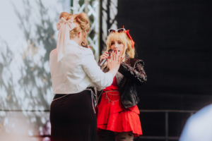 Harmoonics Zwei Künstlerinnen treten auf der Bühne des Elbenwald Festivals auf. Die eine trägt ein schwarzes, mit Nieten besetztes Outfit und ein rotes Kleid, während die andere in einem weißen Oberteil mit einer großen Schleife auf dem Rücken singt.