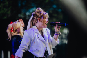 Harmoonics Sängerin auf der Bühne des Elbenwald Festivals, gekleidet in einer modischen weißen Jacke, während sie leidenschaftlich ins Mikrofon singt. Im Hintergrund eine weitere Künstlerin in kostümierter Kleidung, die die Atmosphäre des Festivals unterstreicht.