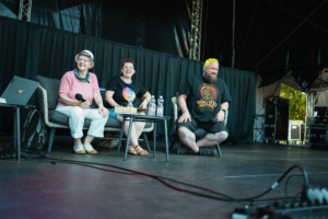 Herr der Ringe Talk - Annika Röttinger, Lisa Kuppler, Tobias M. Eckrich Drei Personen sitzen auf einer Bühne beim Elbenwald Festival, umgeben von Technik und Mikrofonen. Die Frauen tragen bunte Outfits, während der Mann mit buntem Haar lächelt. Im Hintergrund sind Event-Ausrüstungen sichtbar, die die lebendige Atmosphäre des Festivals unterstreichen.