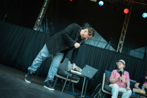 Herr der Ringe Talk - Lisa Kuppler, Stefan Servos Ein Moderator steht auf der Bühne des Elbenwald Festivals, während er ein Mikrofon hält und auf einen Laptop zeigt. Im Hintergrund sitzen Teilnehmer auf einem Sofa, umgeben von einer festlichen Atmosphäre mit bunten Lichtern.