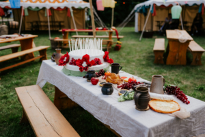 Hobbingen Tisch mit festlicher Dekoration beim Elbenwald Festival, gedeckt mit einer großen Torte, frischem Obst, Backwaren und rustikalen Keramiktassen. Im Hintergrund sind Zelte und Sitzgelegenheiten zu sehen, die eine einladende Atmosphäre schaffen.