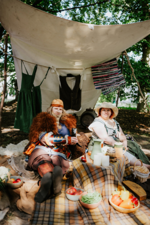 Zwei verkleidete Teilnehmer des Elbenwald Festivals sitzen entspannt unter einem Zelt. Umgeben von rustikalem Geschirr und frischen Lebensmitteln, strahlen sie eine einladende Atmosphäre aus, die den kreativen und gemeinschaftlichen Geist des Festivals widerspiegelt.