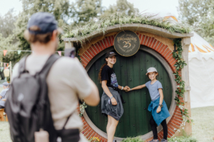 Zwei Kinder posieren vor einer Hobbit-Tür mit einem "Der Herr der Ringe"-Schild beim Elbenwald Festival. Im Hintergrund sind bunte Wimpel und Festivalbesucher zu sehen, die die festliche Atmosphäre unterstreichen.