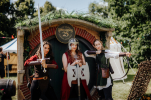 Hobbingen Drei Frauen in fantasievollen Kostümen posieren vor einer Hobbit-Höhle beim Elbenwald Festival. Sie halten ein Schwert, ein Schild und einen Bogen, umgeben von einer lebhaften Festivalatmosphäre. Das Event feiert die 25 Jahre von "Der Herr der Ringe".