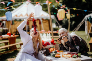 Hobbingen Zwei kostümierte Teilnehmer des Elbenwald Festivals sitzen an einem festlich gedeckten Tisch. Die blonde Person hält eine Traube über ihren Mund, während die andere mit einem Stück Fisch lächelt. Im Hintergrund sind bunte Dekorationen und Festzelte zu sehen.