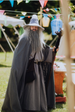 Ein verkleideter Mann mit grauem Umhang und langem Bart hält einen Stock in der Hand. Im Hintergrund sind bunte Wimpel und Zeltstrukturen zu sehen, die die festliche Atmosphäre des Elbenwald Festivals unterstreichen.