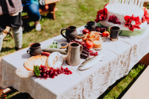 Hobbingen Tisch mit rustikalem Essen und Getränken auf dem Elbenwald Festival. Zu sehen sind verschiedene Speisen wie Brot, Wurst, Obst und ein dekorierter Kuchen, umgeben von Tongefäßen und einer grünen Tischdecke, auf einer Wiese präsentiert.