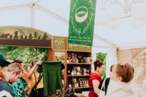 Besucher des Elbenwald Festivals stehen an einem Stand mit Merchandise, darunter ein grünes T-Shirt und ein Banner mit einem bekannten Zitat. Im Hintergrund sind thematische Dekorationen und Produkte aus der Fantasy-Welt zu sehen.