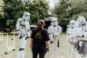 Hobby Horsing Eine Person mit schwarzem T-Shirt steht vor mehreren Stormtroopern, die auf einer Wiese beim Elbenwald Festival stehen. Die Atmosphäre ist lebhaft und kreativ, mit bunten Requisiten und einem nerdigen Flair, das die Festivalbesucher begeistert.