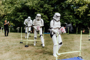 Hobby Horsing Stormtrooper-Kostüme beim Elbenwald Festival, während sie in einer lustigen Herausforderung mit Plüsch-Einhörnern über Hindernisse laufen. Im Hintergrund wird das Event von einem Zuschauer fotografiert. Spaß und Kreativität vereinen sich in dieser einzigartigen Festivalatmosphäre.