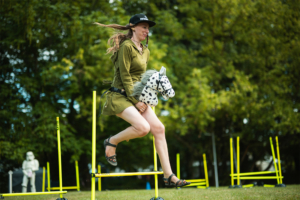 Hobby Horsing Eine Teilnehmerin springt mit einem Plüschpferd über Hindernisse beim Elbenwald Festival, umgeben von einer lebendigen Festivalatmosphäre und grüner Natur. Die Szene fängt den kreativen und verspielten Geist des Events ein.