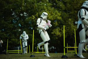 Hobby Horsing Ein Stormtrooper in Kostüm hält ein Plüschpferd und läuft durch einen Hindernisparcours mit gelben Stangen. Im Hintergrund sind weitere Stormtrooper zu sehen, während das Festivalgelände von Bäumen umgeben ist.