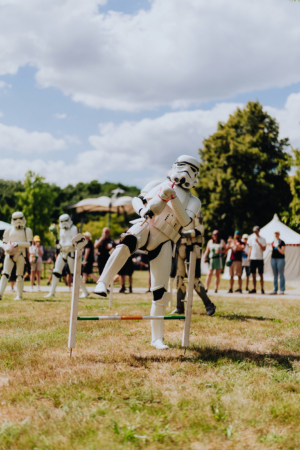 Hobby Horsing Ein Stormtrooper in voller Rüstung springt über ein Hindernis während des Elbenwald Festivals. Im Hintergrund sind weitere Stormtrooper und Festivalbesucher zu sehen, die die unterhaltsame Darbietung beobachten. Sonniger Tag mit blauem Himmel und Wolken.