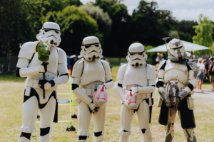 Hobby Horsing Vier Stormtrooper-Kostüme auf dem Elbenwald Festival, halten bunte Spielzeugtiere in der Hand. Die Szene zeigt die kreative Mischung aus Cosplay und Festivalatmosphäre im Freien, umgeben von grüner Natur und anderen Festivalbesuchern.