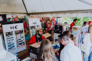 Ichiban Kuji Verkäufer auf dem Elbenwald Festival präsentiert Merchandise und Figuren, während Besucher am Stand interessiert stöbern. Bunte Anime-Poster schmücken den Hintergrund, während Preisschilder von 16 € die angebotenen Produkte kennzeichnen.