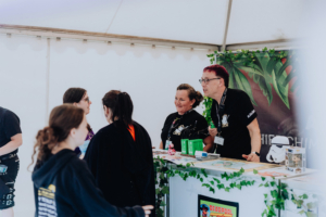 Mitarbeiter am Stand des Elbenwald Festivals interagieren mit Besuchern. Der Stand ist mit Pflanzen dekoriert und bietet Merchandise sowie Informationsmaterial zu Themen aus der Popkultur. Eine freundliche Atmosphäre prägt die Veranstaltung.