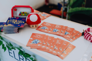 Bunte Broschüren des Elbenwald Festivals liegen auf einem Tisch, umgeben von einem Super Mario Spielzeug und einem roten Pilz. Die Veranstaltung kombiniert Musik und Nerd-Kultur und bietet Besuchern einen Festivalatlas für spannende Erlebnisse.