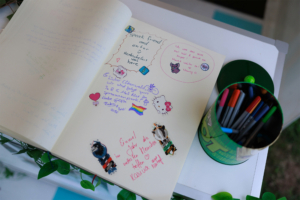 Ein offenes Notizbuch mit bunten Zeichnungen und persönlichen Nachrichten von Besuchern des Elbenwald Festivals, umgeben von einem Behälter mit Markern. Die Seiten sind mit kreativen Stickern und herzlichen Grüßen dekoriert, die die Gemeinschaft des Festivals widerspiegeln.