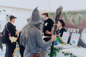 Ein Besucher im Kostüm mit grauem Hut und langem Stab steht an einem Stand auf dem Elbenwald Festival. Im Hintergrund sind Mitarbeiter, die Informationen und Merchandise anbieten, umgeben von grünen Pflanzen und Festivalmaterialien.