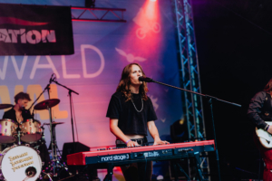 John Leon & Escalation Live-Auftritt beim Elbenwald Festival: Eine Musikerin spielt auf einem roten Keyboard, während im Hintergrund ein Schlagzeuger zu sehen ist. Die Bühne ist mit bunten Lichtern beleuchtet und das Festival-Logo ist im Hintergrund sichtbar.