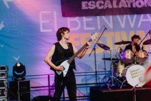 John Leon & Escalation Ein Musiker spielt Bass auf der Bühne des Elbenwald Festivals. Im Hintergrund sind Schlagzeug und eine farbenfrohe Beleuchtung sichtbar, die die energiegeladene Atmosphäre des Festivals unterstreichen.