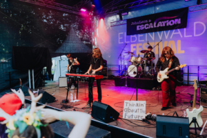 John Leon & Escalation Live-Auftritt der Band JohnLeon & Escalation auf der Bühne des Elbenwald Festivals, umgeben von bunten Lichtern und einer energiegeladenen Atmosphäre, während das Publikum begeistert zuschaut.