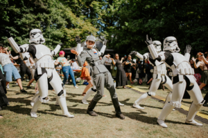 K-Pop-Dance Eine Gruppe von Stormtroopern und einer Person in einem Imperialen Outfit tanzen fröhlich auf dem Elbenwald Festival. Die Menge im Hintergrund genießt die Atmosphäre des Festivals, das Musik und Nerdkultur vereint.
