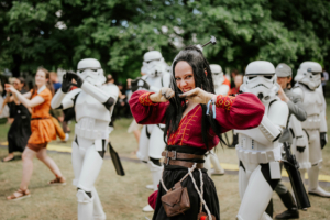 K-Pop-Dance Cosplayer in historischer Kleidung posiert fröhlich im Vordergrund, während Stormtrooper im Hintergrund synchron tanzen. Die Szene spiegelt die lebendige Atmosphäre des Elbenwald Festivals wider, wo Musik und Fantasie aufeinandertreffen.