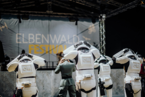 K-Pop-Dance Eine Gruppe von Cosplayern in futuristischen Rüstungen steht auf dem Elbenwald Festival, während sie sich auf die Bühne konzentrieren. Im Hintergrund ist das Festival-Logo sichtbar, das die einzigartige Mischung aus Musik und Nerd-Kultur repräsentiert.